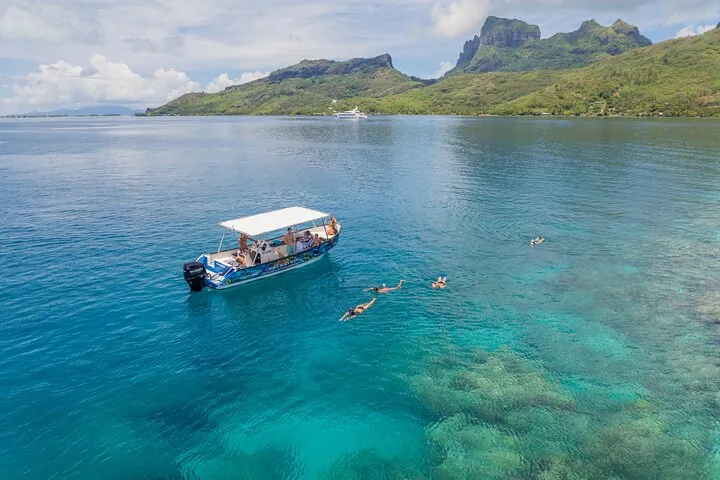 Boat anchored below green peaks with snorkelers swimming over coral
