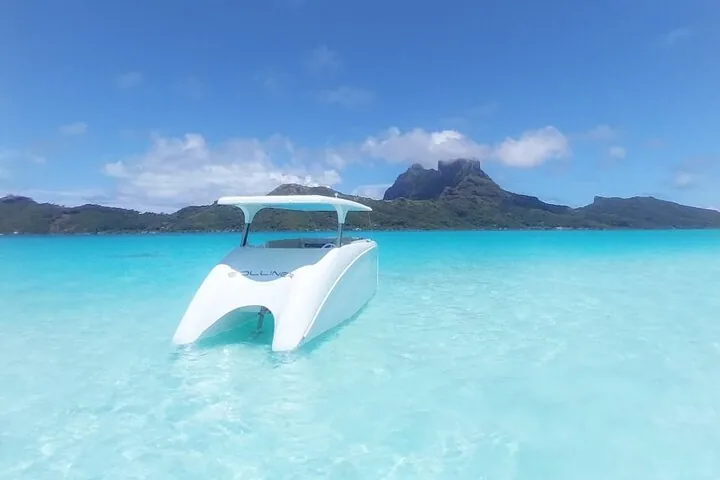 White solar catamaran floating in shallow turquoise water near the island